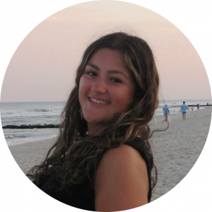 A headshot of Amelia DePino. She is looking over her shoulder at the camera and smiling. There is a beach in the background